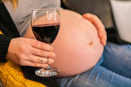 Closeup shot of a glass of red wine in hand of a woman in later stages of pregnancy, with swollen baby bump blurry in background, risk to unborn childの写真素材