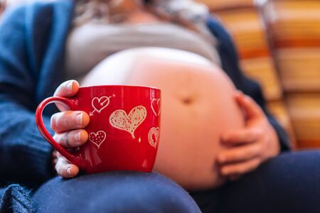 A low angle selective focus shot of a heavily pregnant woman enjoying a hot brew in novelty red...