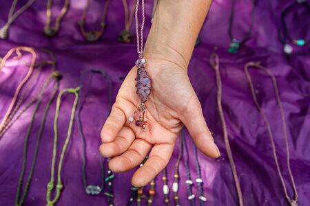 Mans hand holding a handmade necklace with a purple gem or stone. Farmers market jewelery stall with purple cloth decoration in the background.の写真素材