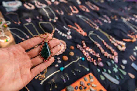 Mans hand holding a dark green stone pendant at a hand made jewelery stand. Farmers market vendor,...