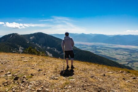 A young man holding a crutch and hiking. A beautiful hilltop landscape. Extremal hiker looking at the beautiful scenic viewの写真素材