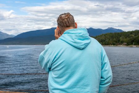 Corpulent middle aged man shot from behind. Passenger standing on the deck of a ferry, looking at the lake and touching his ear with his hand.の写真素材