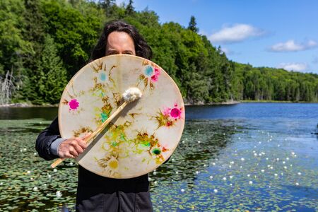 Closeup of long haired native american young man holding sacred native frame drum with fur covered stick at lake with waterlilies covering faceの写真素材