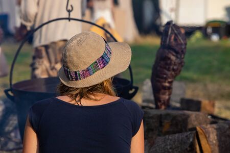 A woman wearing a brimmed hessian hat is seen from the back, sitting by a cauldron cast iron pot...