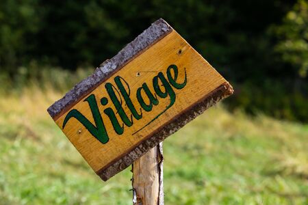 A close up selective focus shot of a skewed handmade wooden sign saying village, fixed to a wood pole at a campsite with blurry green backgroundの写真素材