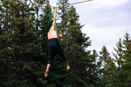 A close up view of a strong man hanging by one hand from a tightrope wire during a gymnastic...