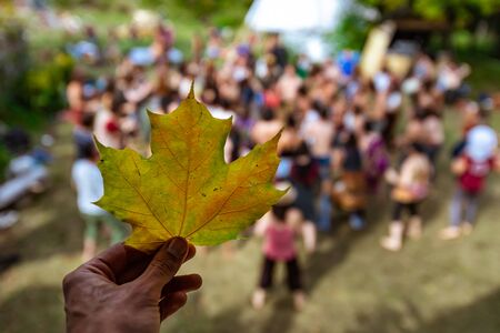 A first person selective focus view of a hand holding a green and yellow leaf near a large group of people, standing in a forest clearing at earth festivalの写真素材
