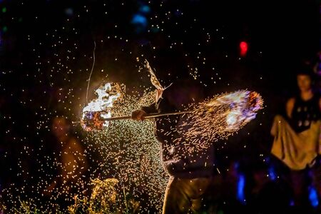 Glowing sparks and embers are seen flying from a lance during a fire dance performance as a man uses flaming pole, with blurry onlookers in backgroundの写真素材