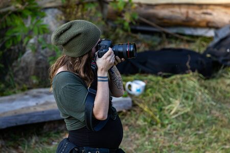 A high angle and side profile view of a professional female photographer, with tattooed arms and wearing a green beanie hat, taking photos in natureの写真素材