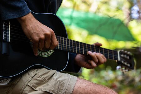 Close up and side view of a talented man, wearing shorts at a campsite during an earth festival, playing guitar with natural green bokeh in backgroundの写真素材