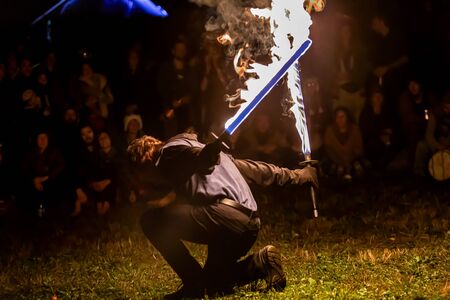 A skilled fire dance performer is seen with two flaming swords outdoors by night, with blurry...