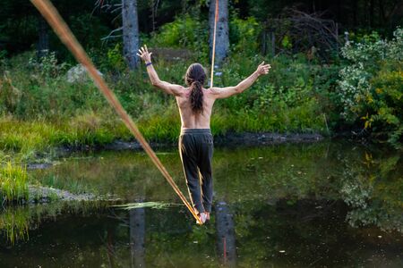 A male slack wire artist is seen practicing slacklining over a water pond in nature, balancing on rope anchored between two trees in woodland with copy spaceの写真素材