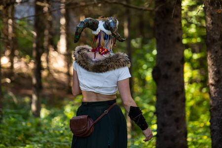 Caucasian woman is seen from the back in selective focus, wearing magical witchcraft horns, horned goddess costume at multicultural festival in forestの写真素材