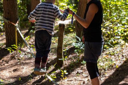 A caring mother is seen teaching a young boy how to balance on a slackline, anchored between two...
