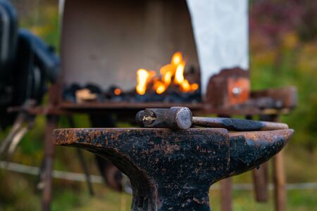 A close up selective focus shot of an old fashioned metalwork anvil and mallet with blurry flaming furnace in background during metalsmith demoの写真素材