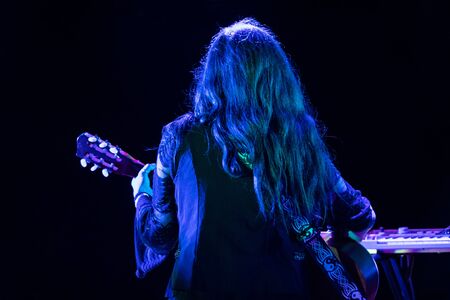 A female folk musician is seen from the back, playing a guitar during a music set with blue stage...
