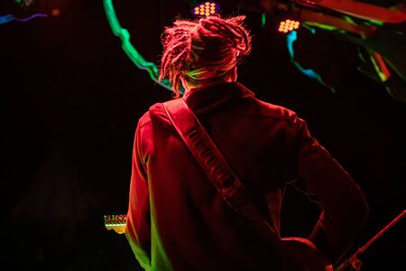 A bohemian man is seen from behind with dreadlocks, playing a guitar on stage with shallow depth of field against a black background by night, copy space to leftの写真素材