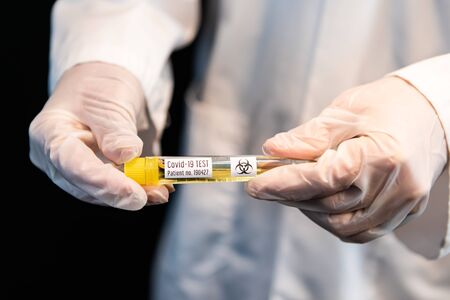 Selective focus closeup of vial with yellow liquid labeld covid-19 test held by gloved hands in...