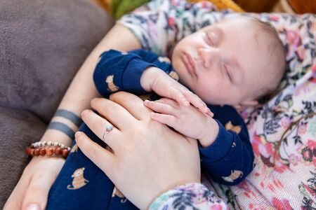 Selective focus shot of young infant boy snoozing in the arms of his mother, her hand adorned with a...