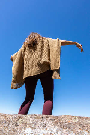 Rear view from below of a woman in a brown crochet poncho sweater against a bright blue sky,dancing a ritual, tribal energy dance.の写真素材