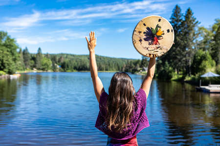 Young woman acting a ritual dance on the shore of a Canadian lake holding a native leather hand painted drum or tambourine in her raised hands.の写真素材