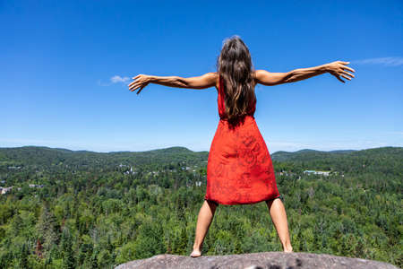 View from behind of a young woman in a red dress opening her arms wide and raising her head to the sky in front of a canadian mountain landscape.の写真素材