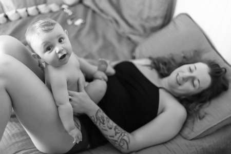 Mother and baby son cuddles. Baby sitting on his mothers belly and looking at camera with a funny, excited expression. Black and white.の写真素材