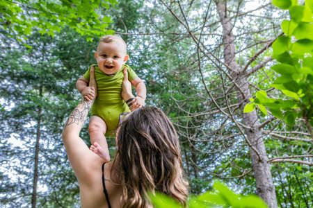Portrait of a mother and her baby, outdoor in the woods. Mommy is holding her son in her arms up high while baby laughs adorably looking at camera.の写真素材