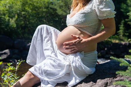 Close up of the naked belly of a pregnant young woman dressed in white, sitting on a big rock next to a river and touching her belly.の写真素材