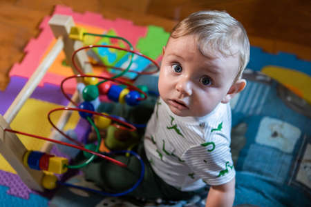 Portrait of creative little curious baby boy sitting on small cushion in living room playing with...