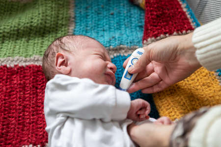 Unwell newborn baby lying on crocket knitted bed crying while selective focus of hands of young female holding thermometer to check temperatureの写真素材