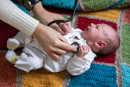 Selective focus of hands of young female doctor holding newborn crying baby hands on knitted bed and checking health using stethoscopeの写真素材