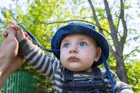 Curious and scared little baby boy wearing hat and casual clothing enjoying in outdoor garden and sitting while holding father hand for supportの写真素材