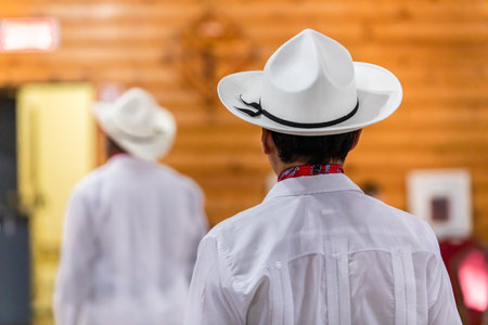 Rear view of young male in white dress costume, with hat and black ribbon tied while standing in room during Dia De Los Muertos in Mexicoの写真素材