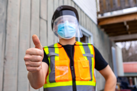 selective focus. Close up of man with orange and yellow fluo visibility jacket, covid protective face mask and plastic visor giving the thumbs up signの写真素材
