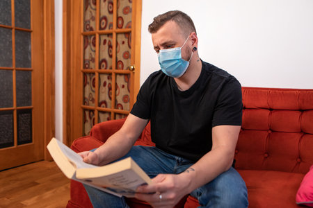 Exaggerated covid prevention. Man reading a book on a red couch, wearing a protective sanitary mask...