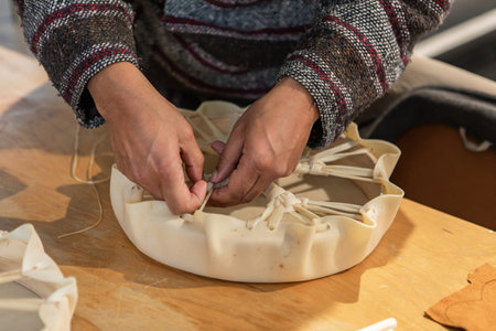 Close up from above of mans hands tying the ends of a piece of leather around a wooden support to create a tambourine. Native drum workshop.の写真素材