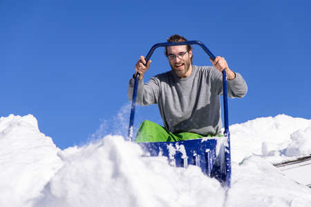 Shot from below of a young man posing with a blue manual shovel from the roof of a house where he is...