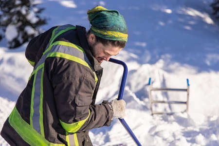 Young man in fluo visibility clothes and blue manual shovel removing knee deep fresh snow from a country house in canada during a sunny winter day.の写真素材