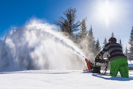 Wide angle view from behind of a man maneuvering a snowplough in the knee deep fresh snow, with a high jet exiting the machine like a snowstormの写真素材