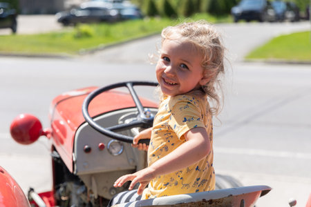 Little kid with blonde hair is sitting on a red and gray tractor, gripping the steering wheel and smiling. Selective focusの写真素材