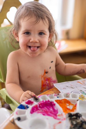 Happy toddler girl covered with colorful paint is sitting at a table and playing with a paint palette. Selective focusの写真素材