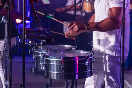 Musician wearing white is playing a set of timbales drums on stage with purple lighting during a...