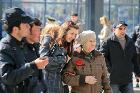 Novosibirsk, Russia - May 9, 2012. Holiday Victory Day. Granddaughter with grandmother, home front worker at the celebration. An old woman has a carnation flower in her pocket. Men walk past.のeditorial素材