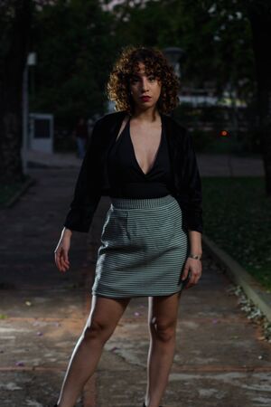 Young girl with curly brown hair, Caucasian skin, red lipstick, black suit and white and black checkered skirt, strolling through the square in the late afternoon and early evening.の写真素材