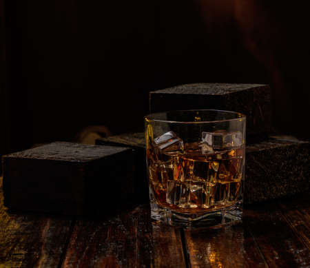 Black varnished wooden table, with a whiskey glass with ice, black background with smoke, yellow lightの写真素材