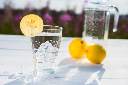 Glass of ice and water decorated with a slice of lemon standing on a white table against a decanter with water and two lemons. Beautiful still life of soft drinksの写真素材