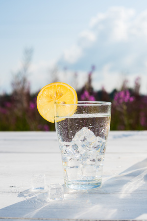 Glass of ice and water decorated with a slice of lemon standing on a white table against a decanter with water and two lemons. Beautiful still life of soft drinksの写真素材