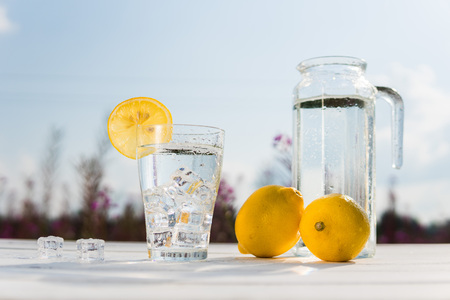 Glass of ice and water decorated with a slice of lemon standing on a white table against a decanter with water and two lemons. Beautiful still life of soft drinksの写真素材