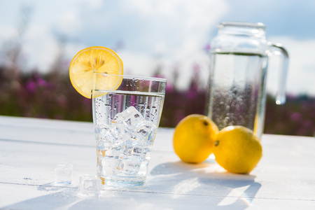Glass of ice and water decorated with a slice of lemon standing on a white table against a decanter with water and two lemons. Beautiful still life of soft drinksの写真素材